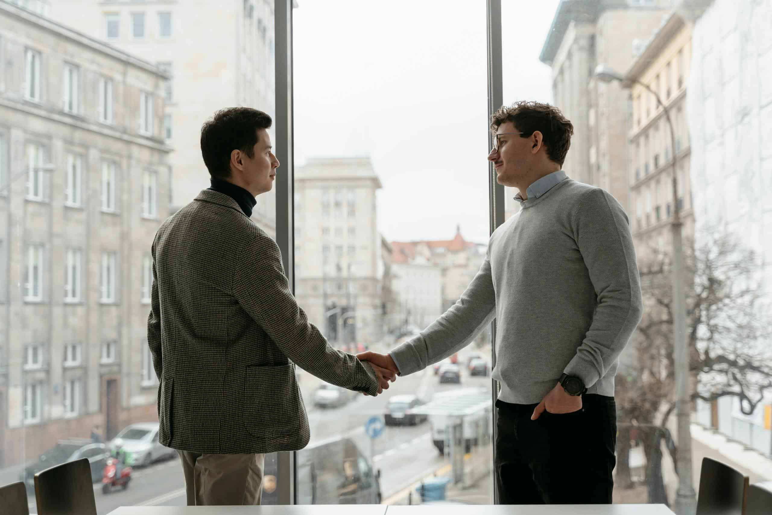 Two businessmen shaking hands in an urban office with city view through large windows.